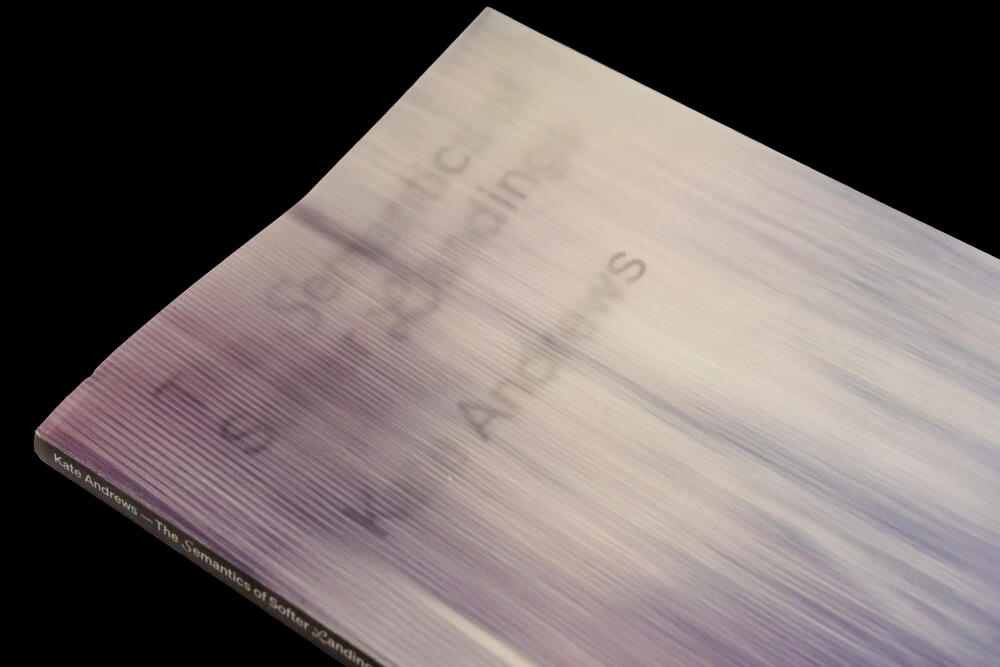 A close-up view of an open exhibition catalog with blurred text visible on white pages, photographed at an angle against a black background.