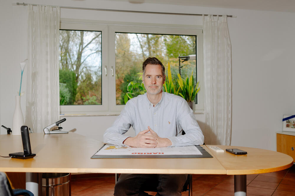 Christian Dürr sits at a wooden desk in an office, wearing a light blue shirt with his hands clasped in front of him. Behind him is a large window with white curtains showing green outdoor scenery. The desk is organized with a laptop, documents, and office supplies.
