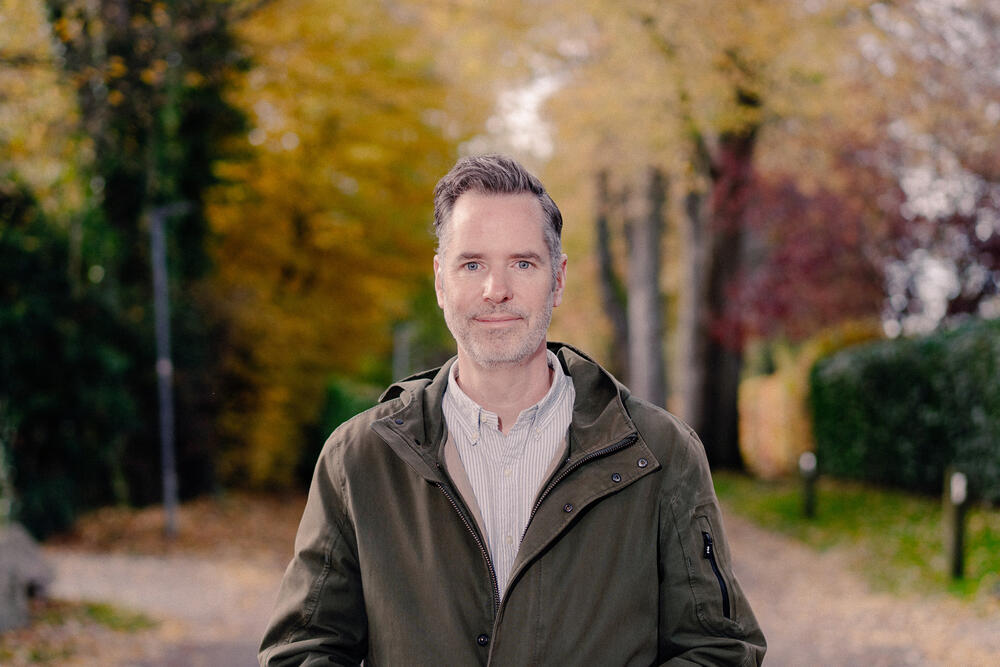 A man with light brown hair wearing a dark olive jacket over a light-colored shirt stands on a tree-lined path during autumn. He faces the camera with a slight smile, with blurred golden and reddish foliage visible in the background.